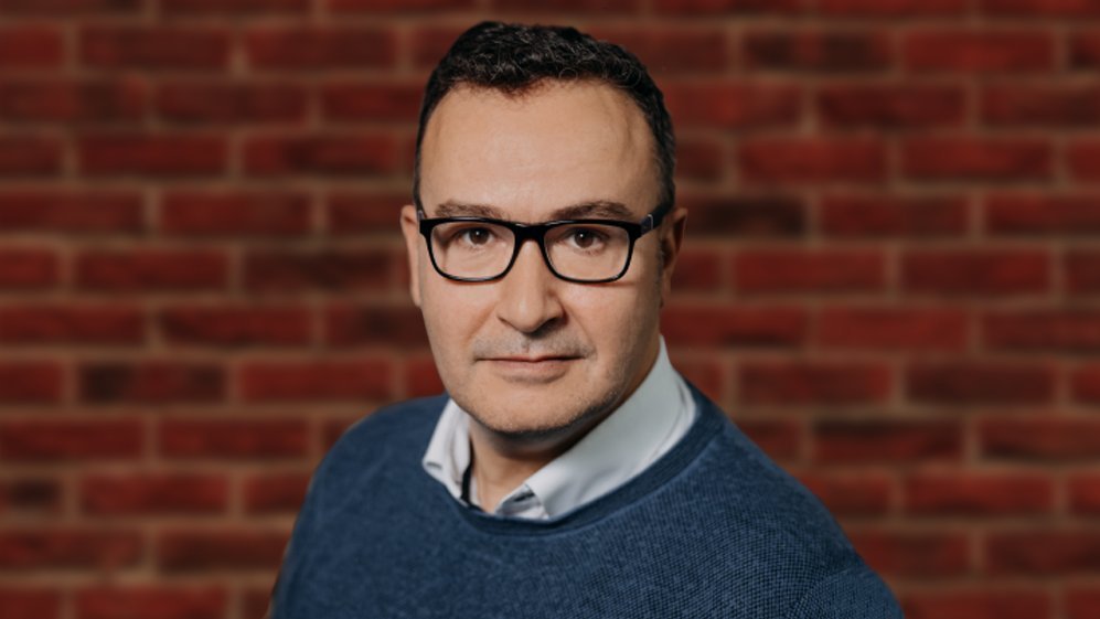 Daniel Abbou, ein Mann mit Brille, steht vor einer Backsteinmauer.